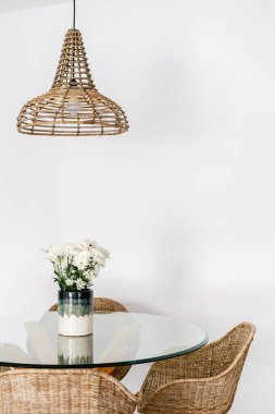Mutfaktaki cam masanın yanında bambu malzemeli hasır koltuğun dikey görüntüsü. Tavan lambası ve iskemle. Boho tarzı odalı rahat bir dairede. Ev dekorasyonu, vazoda taze çiçekler.