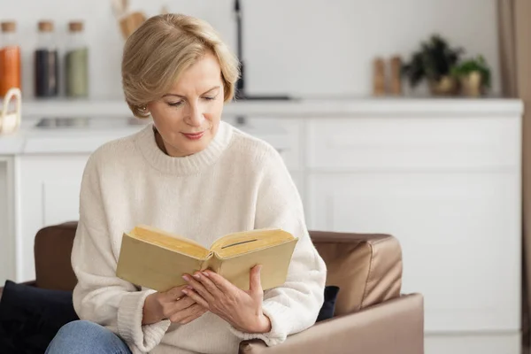 Calm mature woman reading paper book, spending weekend day at home. Smiling senior female sitting in comfortable armchair, enjoying her hobby. Recreation, relaxation concept