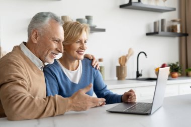 Mutlu, olgun bir çiftin birlikte mutfakta oturup bilgisayardaki video konuşmalarının yan görüntüsü. Karı koca hafta sonunu evde geçirir, internet üzerinden akrabalarıyla iletişim kurarlar.