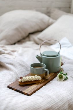 Parlak yatak odasının bir kısmı boho şıklığında, fincanda kahve, çaydanlıkta içecek ve kesme tahtasında ekinlerle. Sabah ışığında kahvaltı tepsisi konfor yatağında duruyor. Hafta sonu konseptiyle evde kal.
