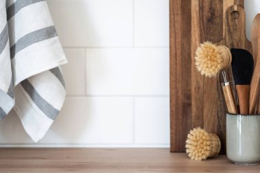 Bambu bulaşık fırçası, ahşap kaşık ve tezgahta pişirilmiş spatula ile seramik bardağın yakın görüntüsü. Mutfak gereçleri, masa örtüsü ve raflardaki tahta kesme tahtaları, fotokopi alanı olan fayanslı duvarlara karşı.