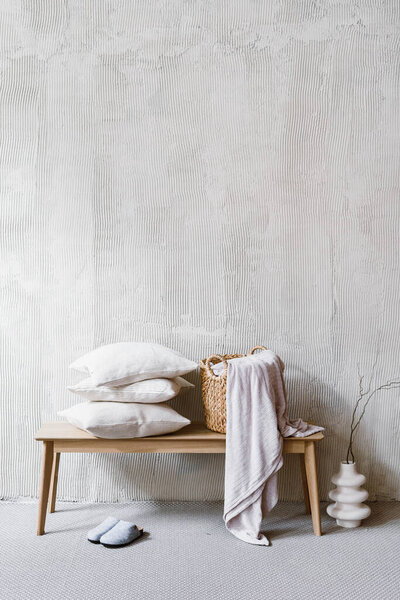 Hotel cleaning service concepts. Vertical shot of pillows, cushions and wicker laundry basket with bedding, duvet or plaid standing on wooden bench in bedroom. Home decor, vase and slippers in room