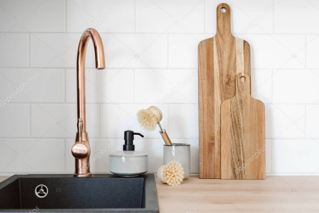 Wooden cutting boards, dishwashing brush in ceramic cup and soap detergent for dishes in dispenser bottle standing near black modern sink with copper faucet. Concept of household objects on kitchen