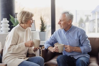 Happy elderly married couple resting at home, talking to each other, drinking hot beverage from coffee or tea mugs. Old friends spending weekend together. Recreation, relaxation concept
