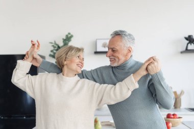 Happy and careless middle aged couple smiling and dancing in kitchen together. Man looking at his grey haired wife with true love, holding her hands, enjoying weekend at home