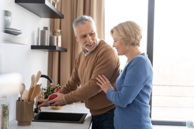 Smiling married couple spending morning, standing on modern kitchen together. Senior man washing fresh red apple and enjoying talk with his lovely wife. Concept of happy family at home