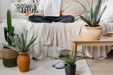 Evde sabah meditasyonunda kadın görüntüsü. Kadın günlük yoga antrenmanı yapıyor, lotus pozisyonunda yatakta oturuyor, Mudra jesti yapıyor ve boho şıklığıyla aydınlık yatak odasında vakit geçiriyor.