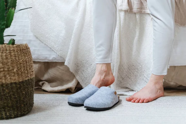 Cropped view of barefoot woman in pajamas pants wearing slippers, standing on rug against cozy bed. Female start her perfect morning at lazy weekend. Part of lady in bright bohemian chic bedroom