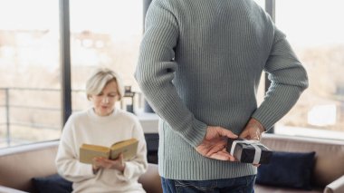 Karısı kanepede oturup kağıt kitap okurken, bir yandan da elinde hediye kutusu olan bir adamın dikiz aynasından kesilmiş görüntüsü. Sürpriz, doğum günü veya yıldönümü konsepti