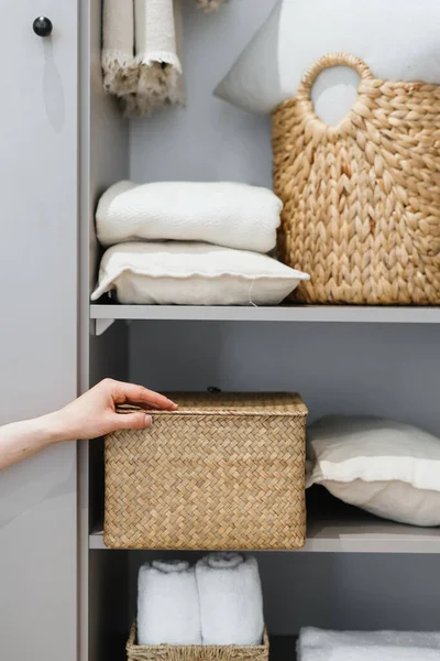Cropped view of neat housewife puts wicker box with clothes on wardrobe shelf, during general cleaning by modern storage system. Concept of comfortable organization of space in apartment