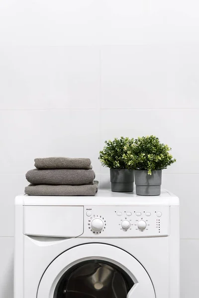 Fresh grey towels and plants in pot on white washing machine in room. Bright apartment interior design. Concept of purity. Laundry idea. Cleaning process. Space for text