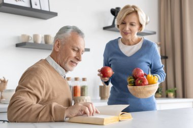 Orta yaşlı bir adam mutfakta karısına kağıt kitap okurken, karısı elinde taze meyve ve sebzelerle kâse tutuyordu. Hafta sonunu birlikte geçirme kavramı. Aile çifti evde iyi vakit geçiriyor.
