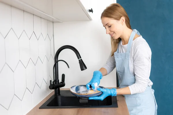 Side view of housewife in apron and gloves doing household duties. Washing plates in sink faucet in kitchen. Kitchenware cleaning in contemporary apartment