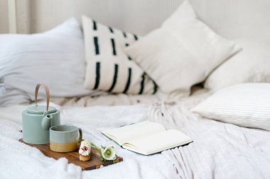 Konfor yatağında kahvaltı tepsisine yakın açık kitap ya da günlük. Kahve fincanı, çaydanlık ve ekinlerin yanında boş sayfalar olan not defteri. Bohem şık yatak odasında mükemmel bir sabah.