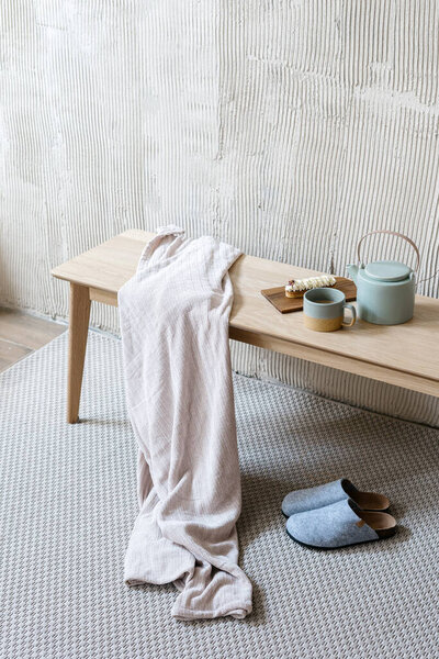 Vertical shot of wooden bench with cozy plaid, eclair on cutting board and teapot with beverage over slippers on rug. High angle view of morning breakfast and drinks on coffee table in bedroom