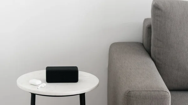 Close up view of portable music speaker with earphones in case lying on white marble table near comfortable grey couch. Audio equipment and digital device on stand