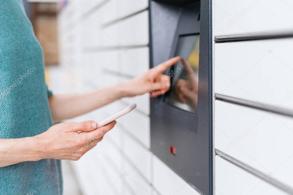 Cropped view of woman standing near automatic post terminal with online app on smartphone, enters the security code on touch screen display and open the deposit locker with delivered parcel