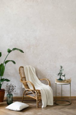 Bamboo armchair with white knitted plaid standing against grey copy space wall in living room. Loft style apartment with boho decor, comfortable rattan chair and potted houseplants