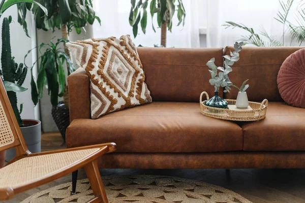 Cozy living room in bohemian apartment with oriental ornament cushions and breakfast tray on comfortable couch. Home decor and eco leather sofa against potted house plants