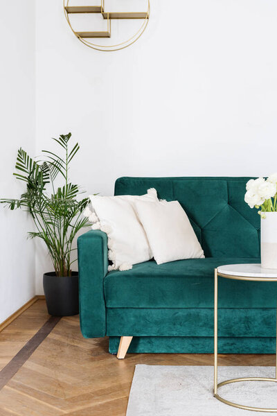 Boho design of living room with emerald velours couch, white cushions, coffee table with flower in ceramic vase and potted houseplant on the floor, closeup