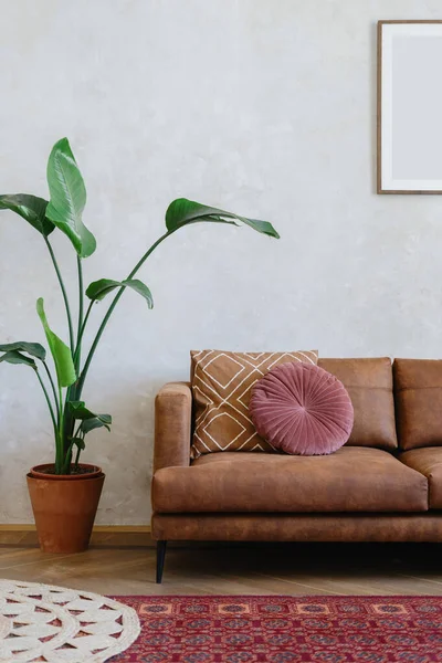 Modern design of living room with brown eco leather couch, soft cushions, mirror on wall, houseplant in clay rustic pot and red carpet on the floor indoors