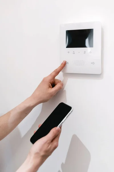 Smart home technology concept. Cropped view of woman with smartphone, controlling temperature in the living room with online app on her phone, choose program on digital panel with touch screen