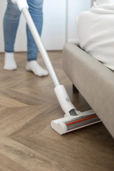 Vertical view of housewife with handheld appliance vacuuming parquet floor under the bed. Concept of wet cleaning in bedroom. Cropped woman using cordless and compact household equipment