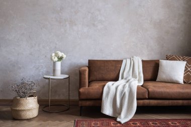Oturma odasının iç tasarımı kahverengi eko deri kanepe, örgü battaniye, yastıklar seramik vazoda çiçek açan yan masa ve yerde hasır sepet.