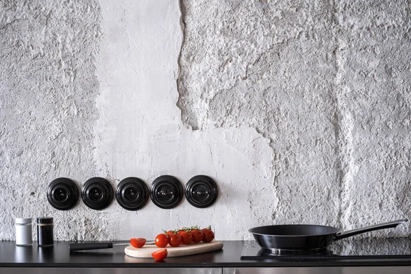 empty frying pan with handle on glass ceramic hob, red fresh tomatoes with knife on wooden board on black worktop at modern kitchen in apartment