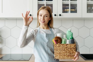 Housemaid from cleaning services holding a box with detergents and household chemical products, showing ok sign, standing on kitchen, looking at camera. Easy and quick housework advertising concept