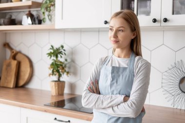 Portrait of lady in apron looking away standing in kitchen. Cleaning service worker after hard work. Rest after work in contemporary apartment