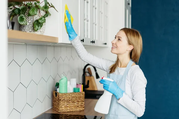 Side view of happy woman in apron and gloves wiping kitchen furniture. Process of removing dirt. Housewife duties. Cleaning service worker job