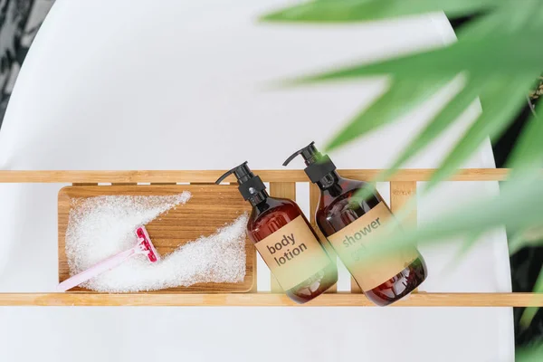 top view of body lotion and shower gel in dispenser bottles, razor and sea salt on wooden caddy on bath or cosmetics products for pampering at home bathtub