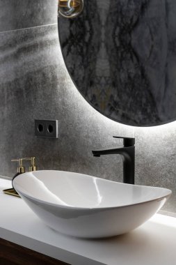 closeup of luxury interior with ceramic washbowl on white countertop, black water tap, grey tile and round mirror on wall in hotel bathroom indoors
