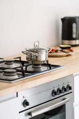 stainless steel cooking pot with handles and glass lid on gas cooker built in wooden countertop and electric oven at contemporary kitchen