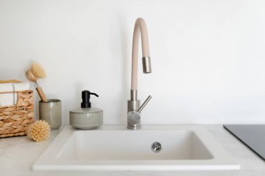 Close up view of new faucet with a water mixer above an empty white sink, next to dishwashing accessory. Organic detergent in soap dispenser bottle, bamboo brush, towel in wicker box on kitchen table