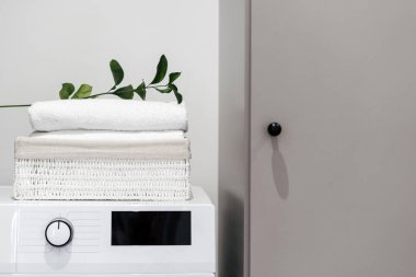 Concept of cleaning service. Green branch over fresh white towels in laundry box lying on top of washing machine with blank screen display. Household equipment, modern appliance in bathroom