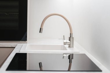 Kitchen faucet above a white empty sink, close to glass ceramic induction stove, against white copy space cabinet with built-in electric oven. Element on modern interior design and household equipment