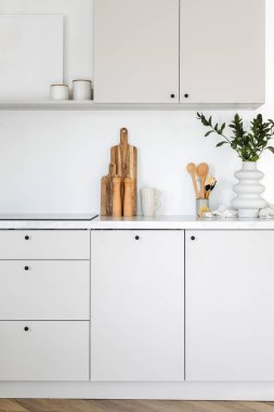 Vertical shot of apartment interior with white kitchen cabinet and cupboards for storing equipment. Tabletop with wooden cutting boards, home decor and kitchenware close to built in induction stove