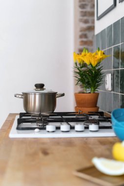 interior of kitchen with gas cooker top built in wooden countertop, stainless steel saucepan with handles and glass lid, yellow blossom flower in pot and green tile on wall