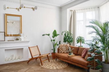Apartment with bright living room in boho style. Interior design with brown couch, new armchair, decorative fireplace with candles, green house plants in clay pots and rug on the wooden floor