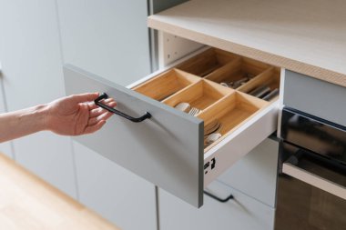 person hand open wooden drawer with metal handle or wood section for keeping silverware, spoons, forks, knife and cutlery in grey cupboard at contemporary kitchen