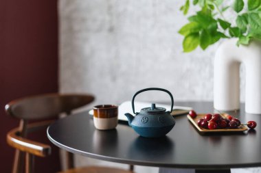 blue clay teapot with brewing chinese tea, ceramic cup and wooden plate with red sweet cherry on dark table at kitchen room or cafe