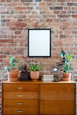 stylish interior design in living room with potted houseplants, books and ceramic statuette decor on wooden cupboard and mockup art poster in black frame on brick wall