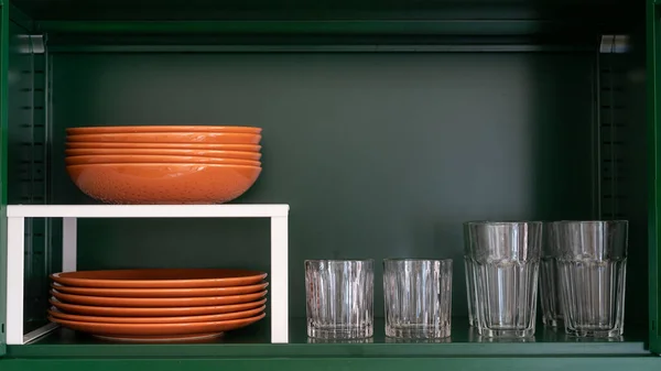 order in kitchen and storage concept, dishware, ceramic plates, brown bowls and glasses on metal green shelf at cabinet