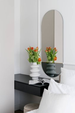 Vertical shoot of bedroom interior with mirror, home decor and nightstand near comfort bed. Hotel room with open magazine and modern ceramic vase with blooming tulips flowers on side table