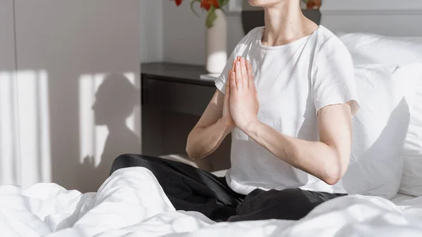 Cropped view of calm woman making praying gesture, sitting on bed in meditation pose. Mental health concept. Recreation, relaxation at morning in comfortable hotel room. Everyday yoga practice