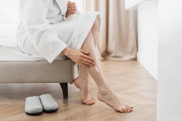 Bodycare, skincare concept. Cropped shoot of woman in bathrobe sitting on bed, applying pampering after shave cream on her legs. Female use moisturizing cosmetics for body, spending morning at home