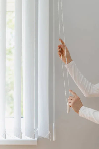 cropped shot of female hands pull rope and open vertical louvers on window in living room at house