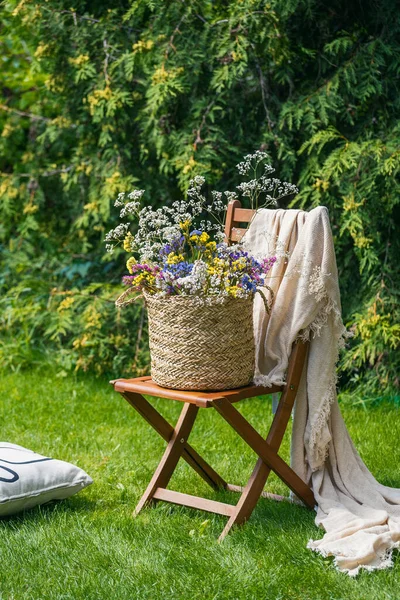 beautiful bouquet of wildflowers in wicker basket on wooden garden seat on green lawn in backyard at summer day